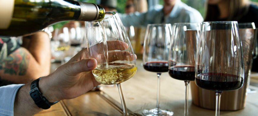 Person pouring white wine into a glass at a lively gathering with multiple wine glasses and joyful people in the background.