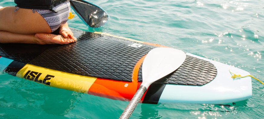 Person sitting on a paddleboard in the water, holding an oar, with close-up view of their lower body and board.