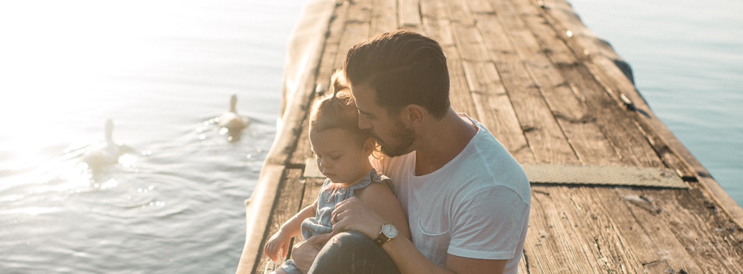 Father and child sitting on a wooden dock, watching swans on a serene lake at sunset.