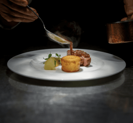 Chef pouring sauce over steak with a fine dining plating, and dark background.