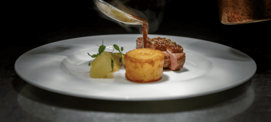 Chef pouring sauce over steak with a fine dining plating, and dark background.